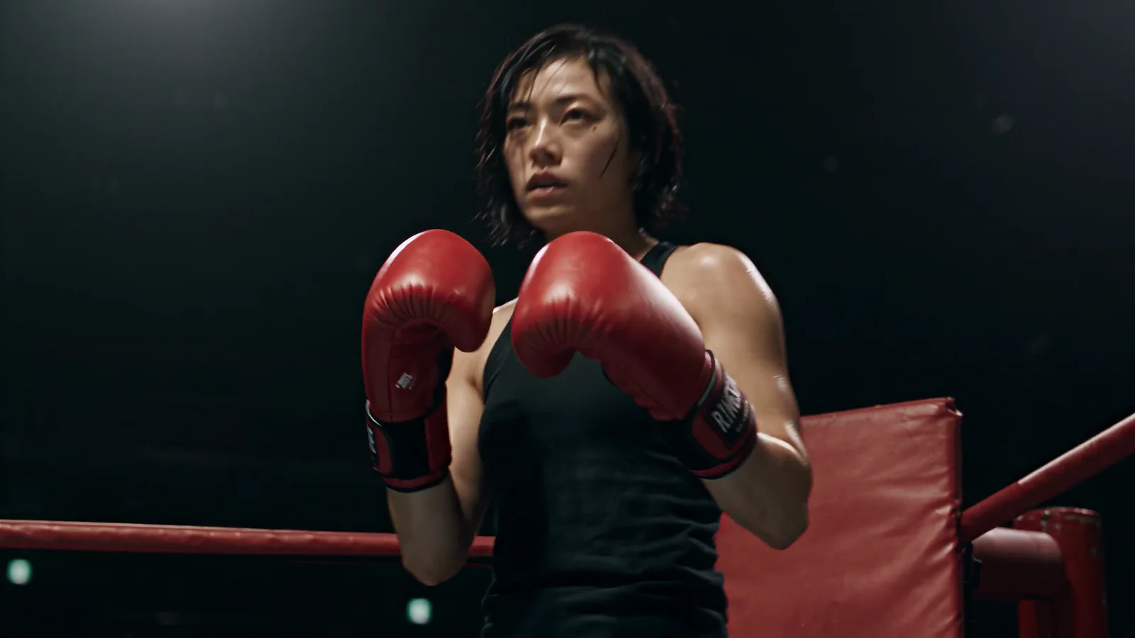 An exhausted East Asian female boxer rises from her corner and brings both red gloves into guard